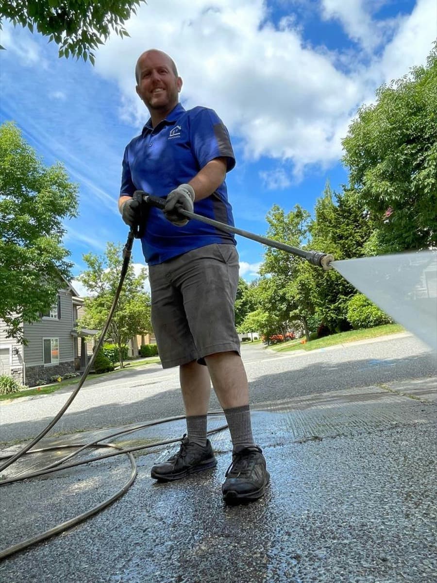 A pressure washing contractor holding a wand with a wide fan-pattern nozzle spraying across a residential driveway