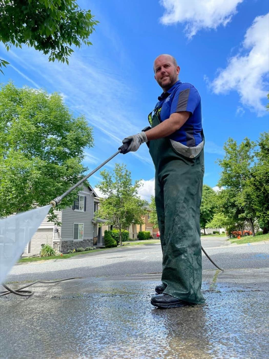 A contractor in chemical-resistant waders applying a wide low-pressure fan of soft wash solution across a residential surface