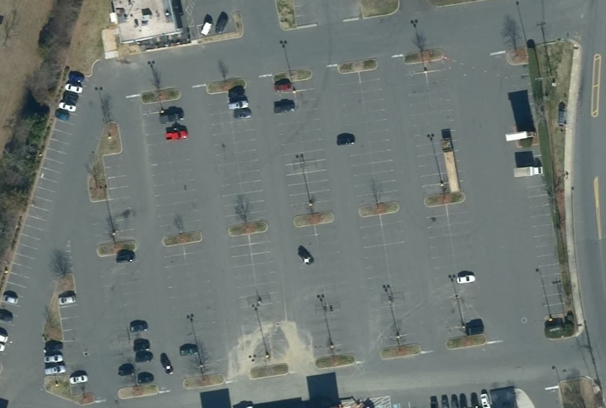 Aerial view straight down on a retail parking field, Feb 2019: Four winters on, the lines are faint ghosts. A pale patch near the bottom.