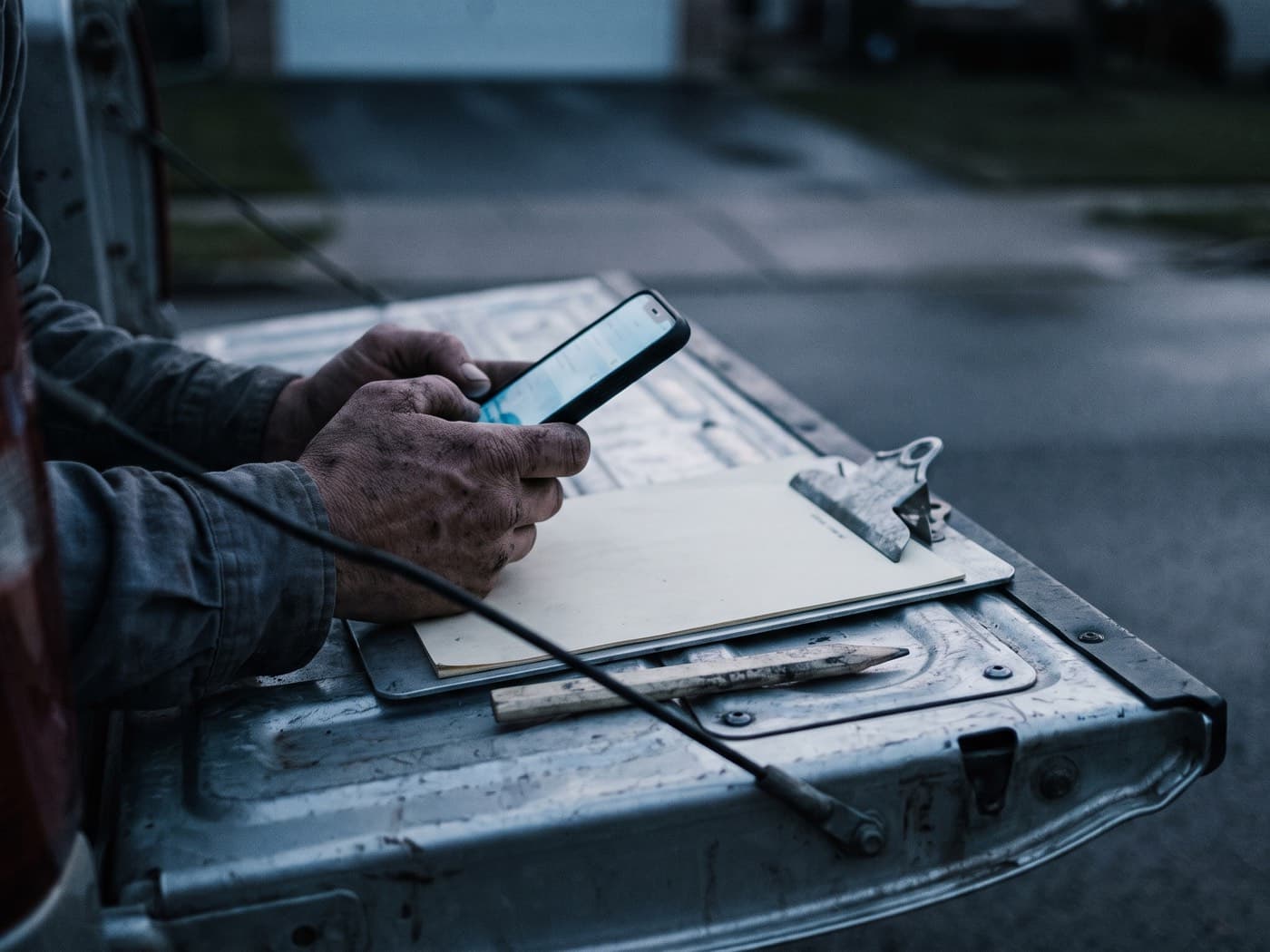 A contractor's hands holding a phone over the open tailgate of a work truck at dawn, beside an aluminium clipboard and a carpenter pencil
