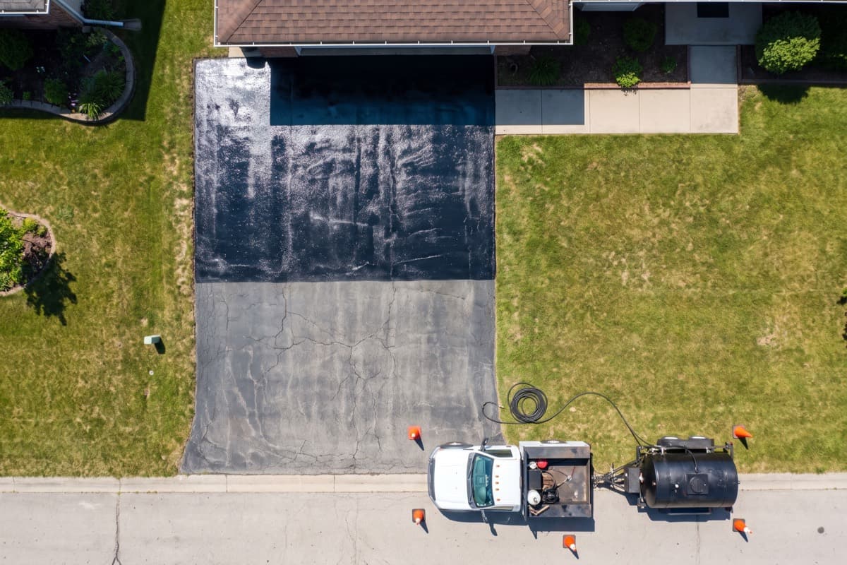 Overhead view of a residential asphalt driveway half-way through sealcoating: the upper half fresh and black, the lower half grey and cracked, with the sealer rig and tank trailer parked at the street