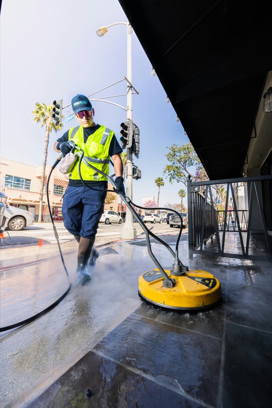 A uniformed contractor walking a surface cleaner along a commercial sidewalk in high-visibility gear