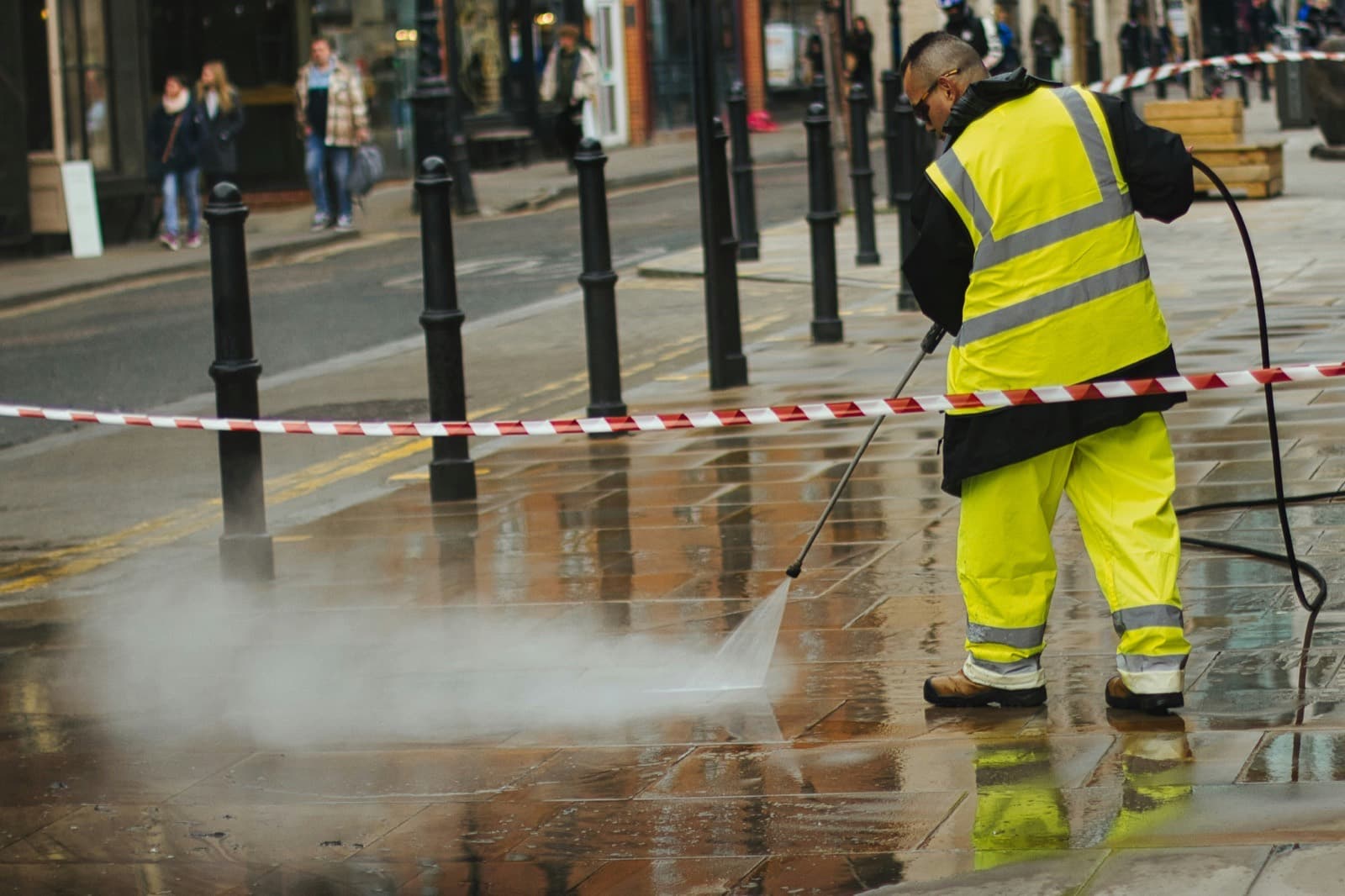 A contractor in high-visibility gear washing a paved public walkway, hose running back down the street