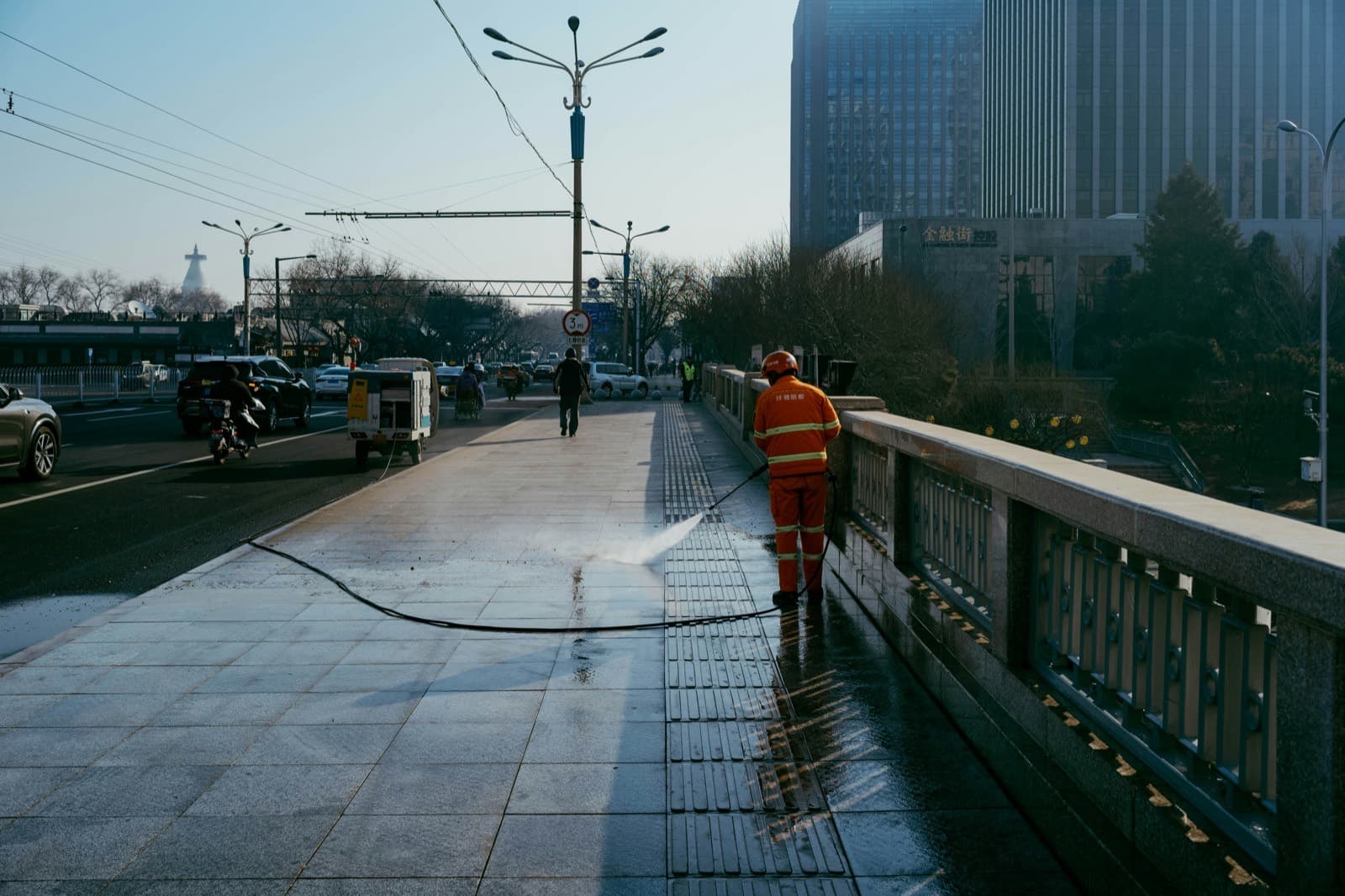 A contractor washing a wide public walkway with the pressure hose running back along the pavement to a wheeled unit parked at the curb