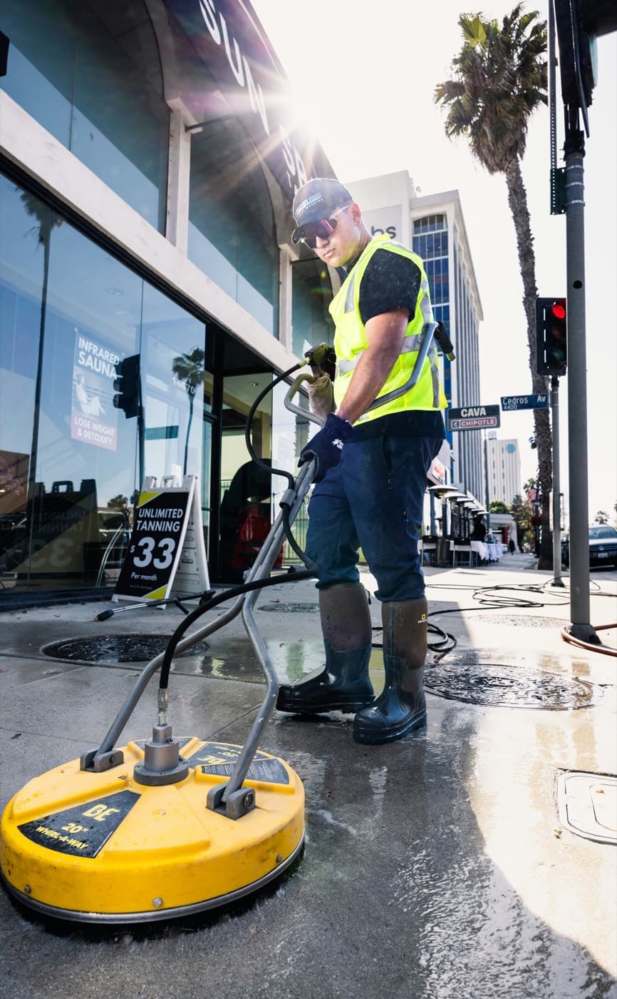 A contractor running a surface cleaner on a commercial street corner in high-visibility gear
