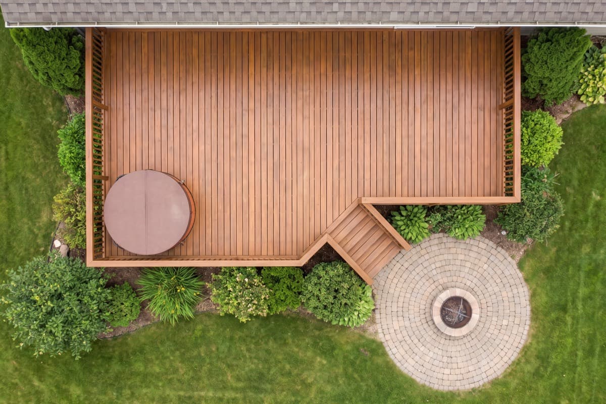 Aerial view of a large stained deck with a hot tub set into one corner, steps down to a circular paver patio with a fire pit at its centre