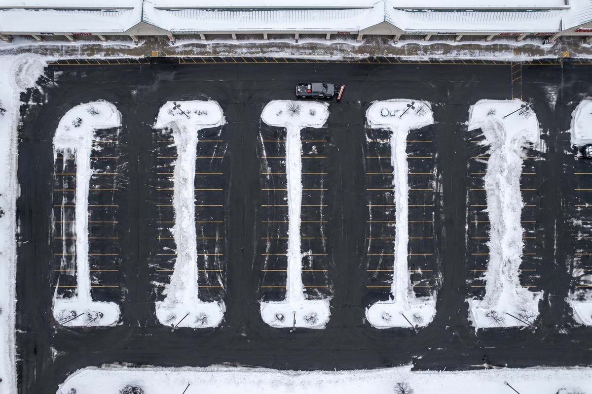 A plowed retail parking lot from above, snow stacked on the islands and a plow truck by the storefronts