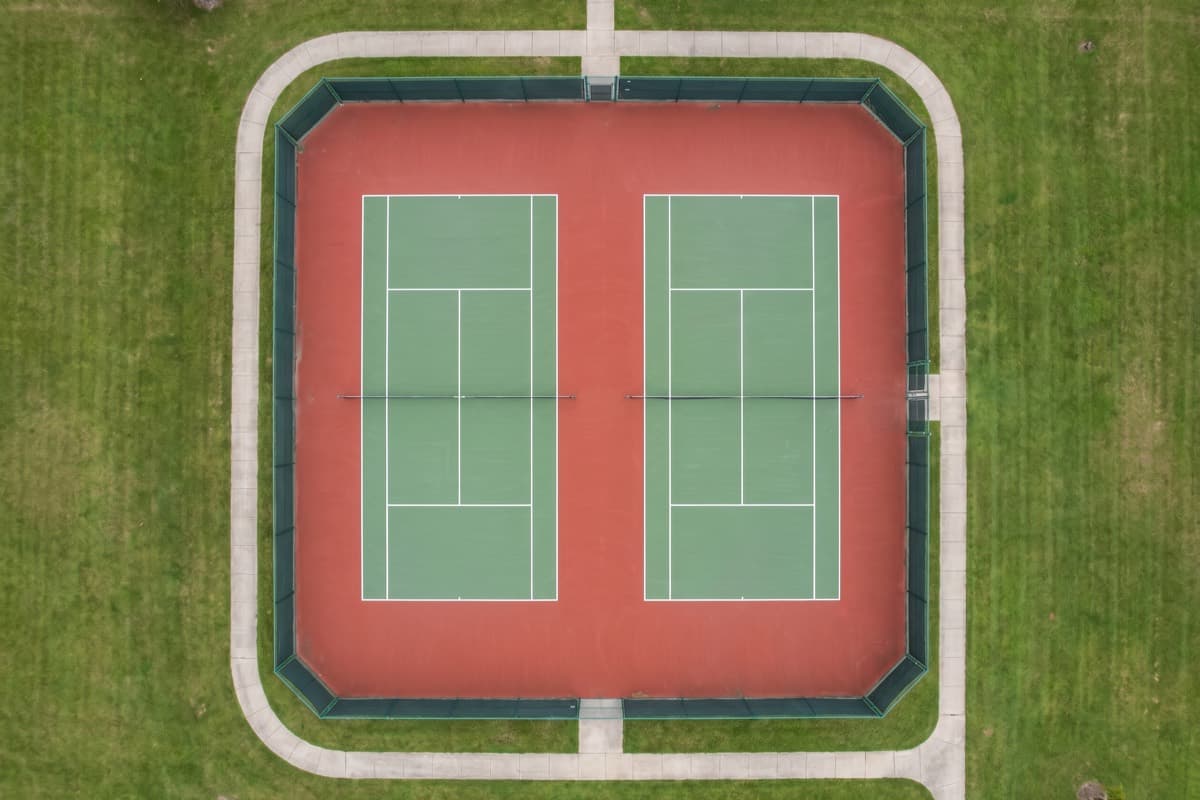 Aerial view of two acrylic-coated tennis courts with white line markings, enclosed by fencing and a perimeter walkway