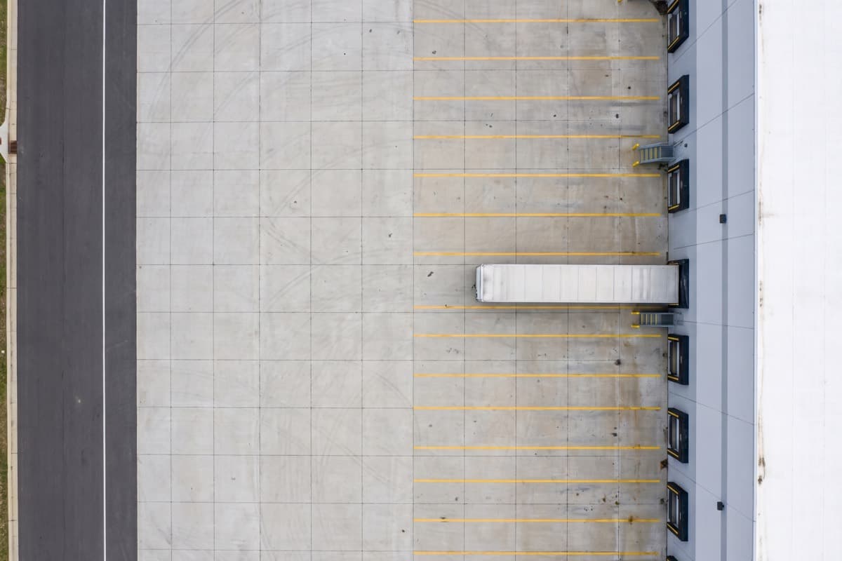 Aerial view of a concrete loading apron behind a warehouse, control joints scoring the slab and yellow dock lines painted between bays